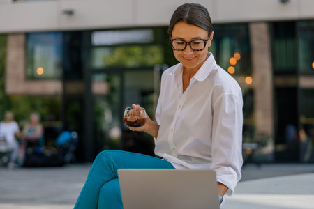 Female manager in eyeglasses drinking coffee and working on laptop on office building backgroundの写真素材
