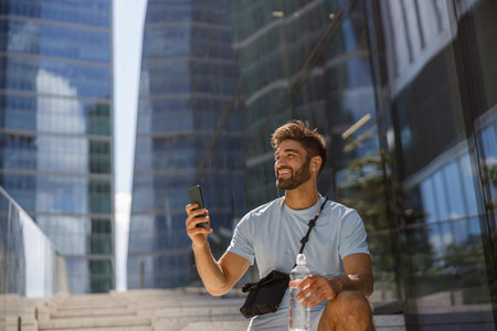 Male tourist with bottle of water resting after walk in modern city and making photo on phoneの写真素材