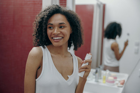 Young smiling woman is holding cosmetics bottle while standing in bathroom and looks awayの写真素材