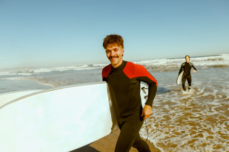 Two happy surfers in wetsuit with his surfboards entering out of sea after surfing on wavesの写真素材