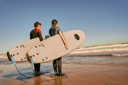 Back view of surfers in wetsuits standing with surfboards and looking on ocean