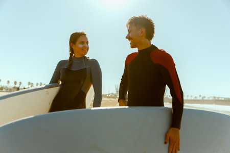 Couple of surfers in wetsuit with surfboards standing on the beach and looking each other with smileの写真素材