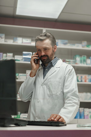 Male pharmacist wearing lab coat working on computer in drugstore and talking phoneの写真素材