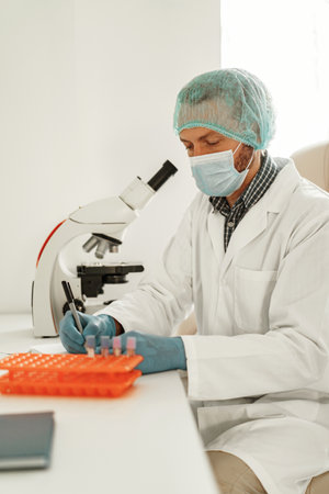 Man scientist in protective mask making notes in report sitting at laboratory with microscope