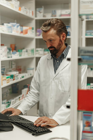 Male pharmacist wearing lab coat working on computer in drugstore