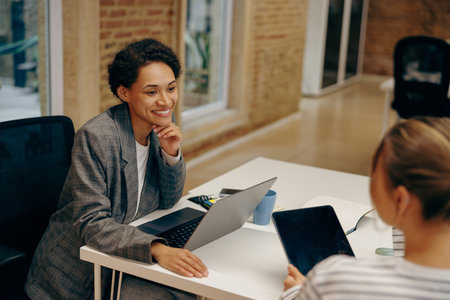 Two stylish smiling female sales managers talking during work while sitting in cozy officeの写真素材