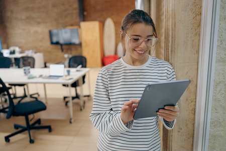 Young smiling female freelancer is working on digital tablet standing on coworking backgroundの写真素材