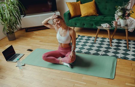 Woman seated on yoga mat in living room on hardwood floor and stretching her neckの写真素材