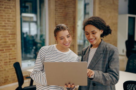 Two busy female coworkers discussing project standing in office with laptop
