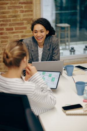 Two smiling female freelancers talking during work on laptop while sitting in cozy coworking