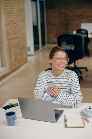 Smiling young business woman working on laptop and use phone while sitting in stylish office