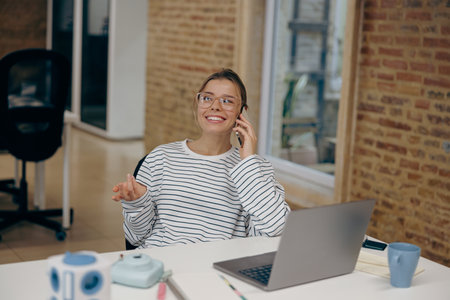 Woman sales manager talking by phone with client sitting in modern coworking and working on laptopの写真素材
