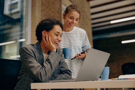 Two smiling female colleagues working on project together and use laptop sitting in officeの写真素材