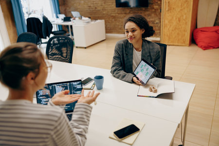 Businesswoman with digital tablet explaining project details to colleague at office deskの写真素材