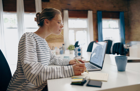 Concentrated female architect working on laptop and making notes while sitting the desk in officeの写真素材