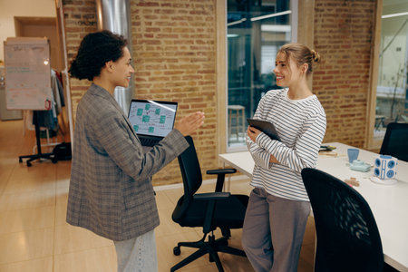 Two smiling female coworkers working on project together and use laptop standing in officeの写真素材