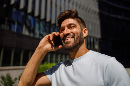 Smiling man in casual clothes talking by phone while standing on city skyscrapers background