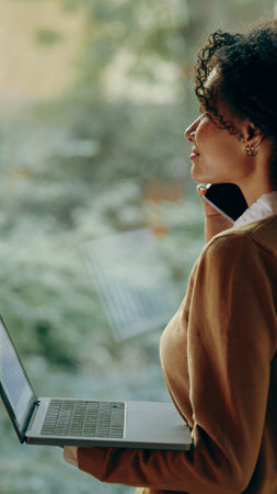 A woman is on a call while efficiently working on her laptop in a cozy and inviting environment
