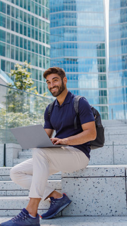 Smiling bearded male freelancer working on laptop sitting on skyscrapers backgroundの写真素材
