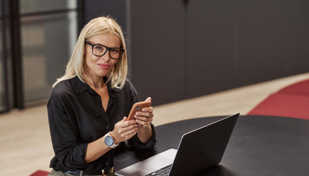 A Professional Woman Actively Engaged in Work Using Her Laptop and Smartphone in an Office Settingの写真素材