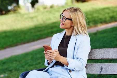 A cheerful, smiling professional woman casually using her smartphone while in a park settingの写真素材