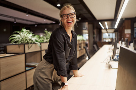 A Smiling Woman Standing Joyfully at a Modern Office Reception Desk, Ready to Welcome Visitorsの写真素材