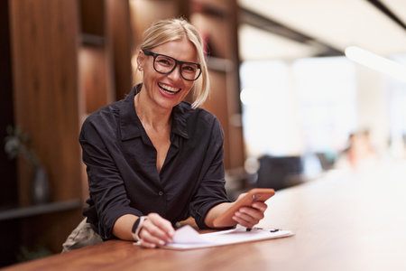 A confident, smiling professional woman sits at her desk, surrounded by a phone and documentsの写真素材