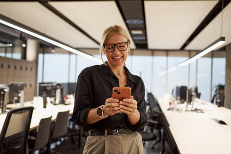 A Smiling Professional Woman Engaging with Her Smartphone in a Modern Office Environmentの写真素材