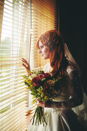 Beautiful red hair bride with flower bouquet standing near window. Image toned in warm colors.の写真素材