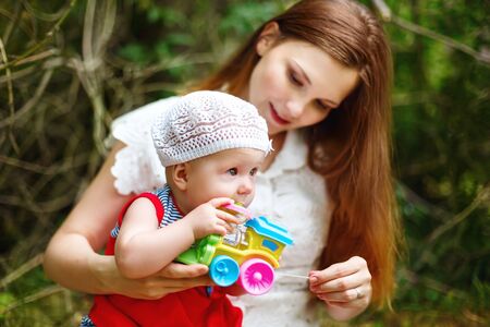 Cute Toddler Baby Girl sitting on Mom`s Hands, playing with Toy. Relaxing in green summer Park. Selective focus on baby`s eyes. Grain added for best impression.の写真素材