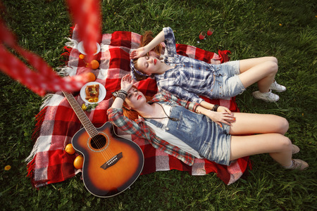 Young Beautiful Smiling Girls Dressed in Pin Up Style Lying and Dreaming on a Red Plaid on Green Grass in Summer Park. Aerial View.の写真素材