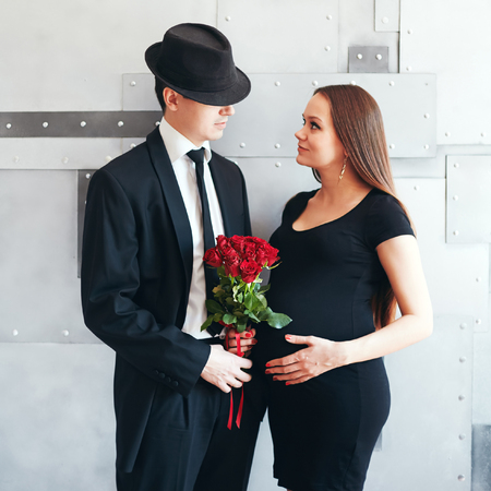 Stylish elegant young couple waiting a baby. Brunette female in black cocktail dress holding bouquet of red roses, man in tuxedo and hat. Looking at each other.の写真素材