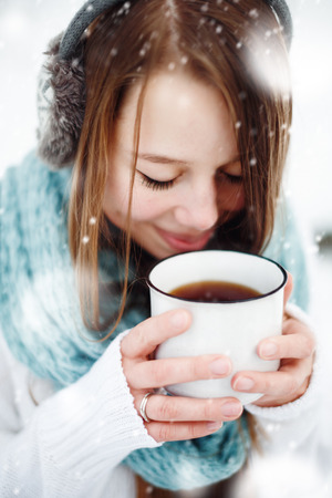 Young Beautiful Female Drinking Hot Drink Outdoors in Winter. Close Up Headshot. Drawn Snow. Selective Focus.の写真素材