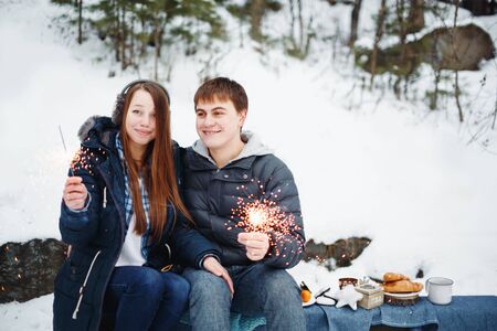 Happy Smiling Couple Sitting on a Log with Holiday Sparklers Celebrating Christmas Outdoors in Winter Forestの写真素材