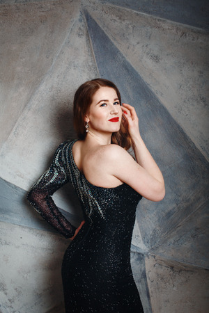 Beautifu Female Model in Luxury Black Dress with Bright Make Up. Woman Standing against Grey Wall.の写真素材