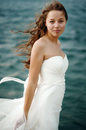 Beautiful Woman in White Dress Standing against Stormy Sea. Blowing Wind Hair. Looking to Camera. Selective Focus. Grain Added for Best Impression.の写真素材