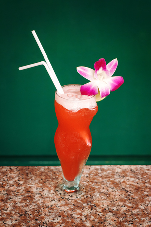 Tropical Red Cocktail with Orchid Flower and Drink Straw on a Table. Funny Glass.の写真素材