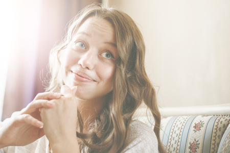 Cute Young Female Headshot. Woman Thinking and Smiling. Big Grey Eyes, Long Curly Hair. Hands near Face. Portrait in Interior. Sun Light Lens Flare.の写真素材