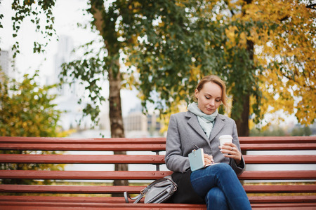 Trendy woman in stylish coat sitting on the bench in city park. Urban scene outdoors. Drinking takeaway coffee and writing or drawing something in notebook.の写真素材