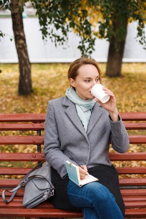 Stylish modern woman in elegant clothes sitting under fall foliage in city park. Urban scene outdoors. Thoughtful and smiling. Drinking takeaway coffee and writing something in notebook.の写真素材