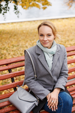 Portrait of young blonde woman in elegant clothes sitting on a bench in autumn park. Urban city scene outdoors. Looking in camera and smiling.の写真素材