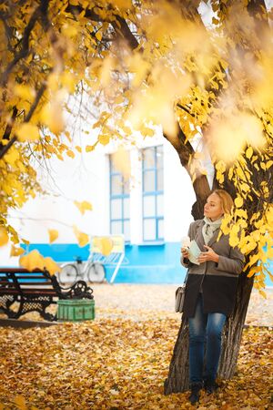 Full body portrait of elegant woman walking with coffee and note in autumn park. Pensive and dreaming. Urban park scene.の写真素材