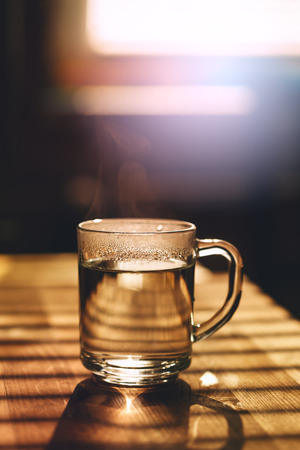 Transparent mug with hot water on kitchen table in sunset light. Image toned, lens flare effect added. Morning concept.の写真素材