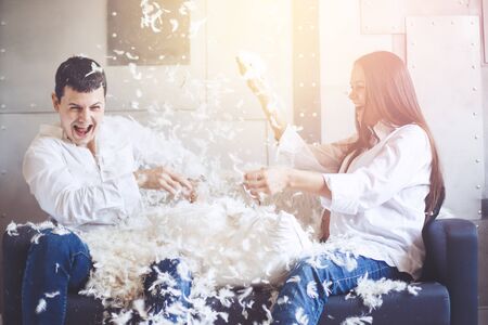 Pillow fight. Young happy pregnant family have fun with fluffy feathers from pillow. Man and woman dressed in jeans and white shirts. Vintage color effect.の写真素材