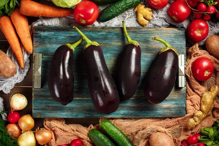 Fresh big eggpants close-up among frame made from ripe vegetables on wooden background. Top view. Aubergines, cabbage, carrot, tomato, onion and other.の写真素材