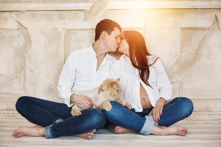 Young family couple with domestic cat waiting a baby. Man and woman in blue jeans and white shirts, kissing. Selective focus.の写真素材