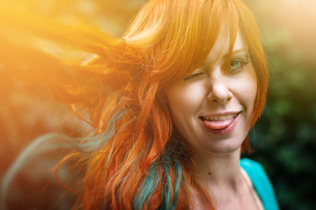 Young trendy freaky woman with colored bright red head having fun and showing tongue. Headshot with sunlight effect, summer mood outdoors.の写真素材