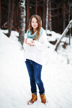 Beautiful woman drinking hot drink in winter park. Winter holidays concept. Full height, looking at camera. Winter landscape on the background.の写真素材