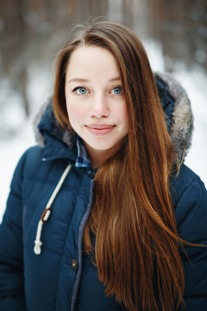 Beautiful woman in winter clothes standing in winter park, smiling, looking at camera. Winter forest landscape on the background.の写真素材