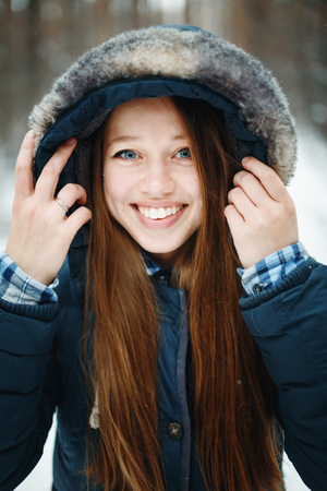 Young smiling woman in winter clothes with hood standing in winter forest, laughing, looking at camera. Winter landscape on the background.の写真素材